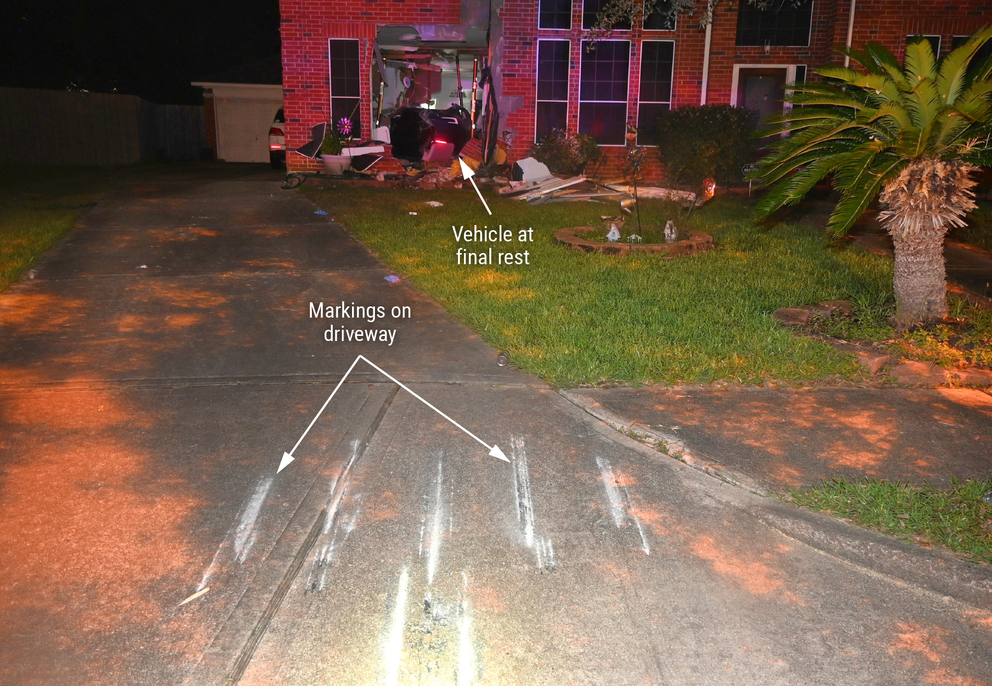 Photograph of the crash location showing markings on the driveway and the vehicle  at final rest inside the residence. 